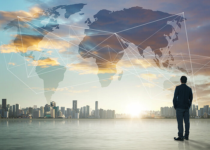 Geschäftsmann schaut auf die entfernte Skyline einer Metropole. Dazwischen befindet sich eine Weltkarte mit Netzwerklinien