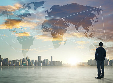 Geschäftsmann schaut auf die entfernte Skyline einer Metropole. Dazwischen befindet sich eine Weltkarte mit Netzwerklinien