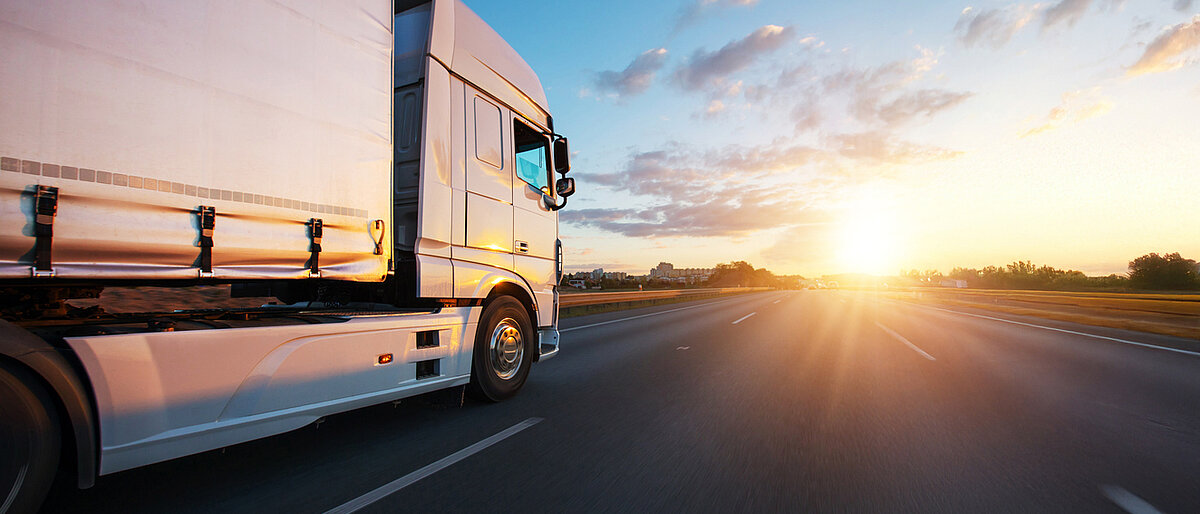Ein LKW fährt auf einer breiten Straße dem Sonnenaufgang entgegen.