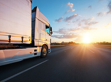 Ein LKW fährt auf einer breiten Straße dem Sonnenaufgang entgegen.