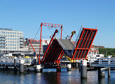 Skyline der Stadt München mit Blick auf Frauenkirche auf der linken Seite, Skyline der Stadt Kiel mit Blick auf eine hochgeklappte Brücke auf der rechten Seite