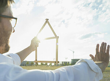 Mann blickt in die Ferne, mit der linken Hand hält er ein zu einem Haus geformten Zollstock