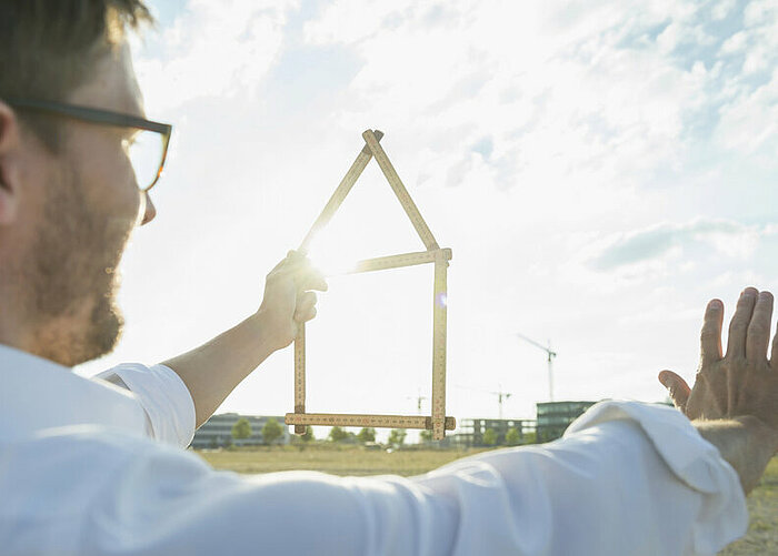 Mann blickt in die Ferne, mit der linken Hand hält er ein zu einem Haus geformten Zollstock