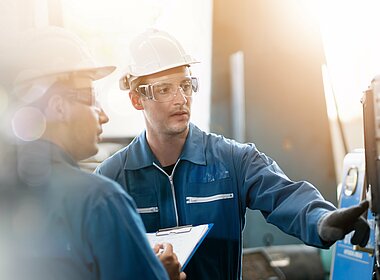 Zwei Arbeiter in Arbeitsmontur mit Helm schauen sich eine Maschine an